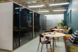 Moderne kantoorlounge aan de Johan Huizingalaan met vergaderpods, ronde tafels, kleurrijke kussens en groen decor.